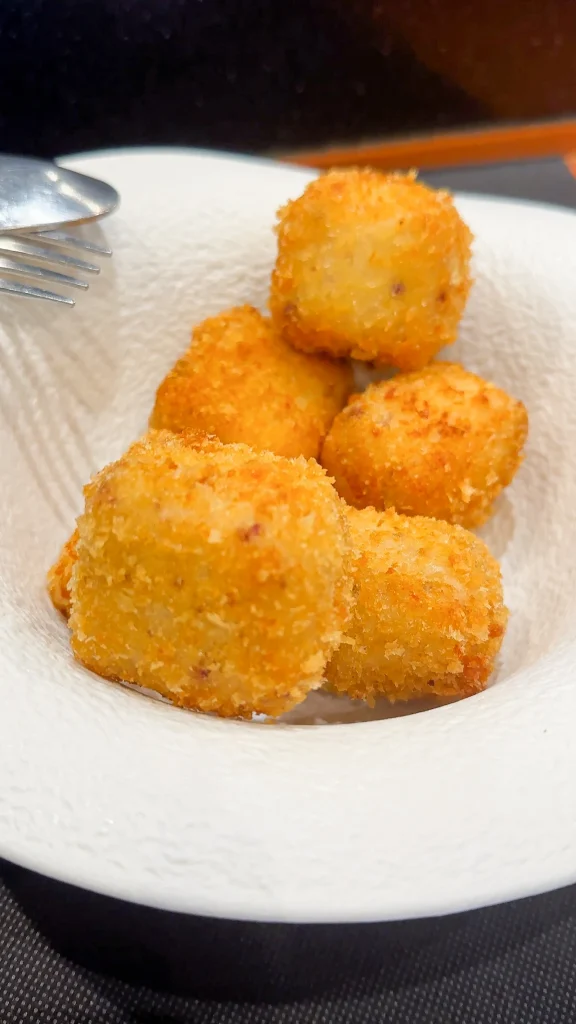 Croquetas de jamón — five round golden-fried croquettes with coarse breadcrumb crust on a white textured plate, Asturias