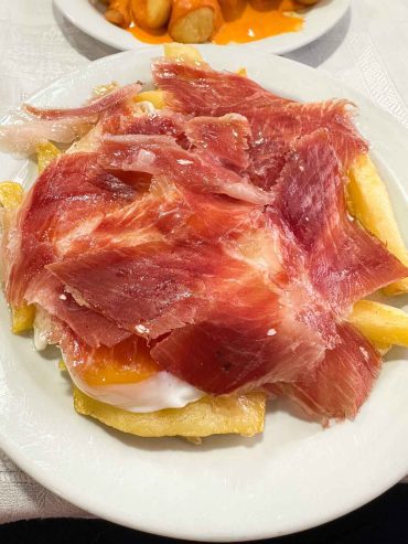 Huevos rotos con jamón — fried potatoes topped with broken egg yolk and draped with jamón ibérico slices on a white plate, Oviedo