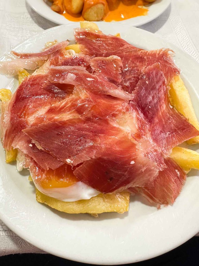 Huevos rotos con jamón — fried potatoes topped with broken egg yolk and draped with jamón ibérico slices on a white plate, Oviedo