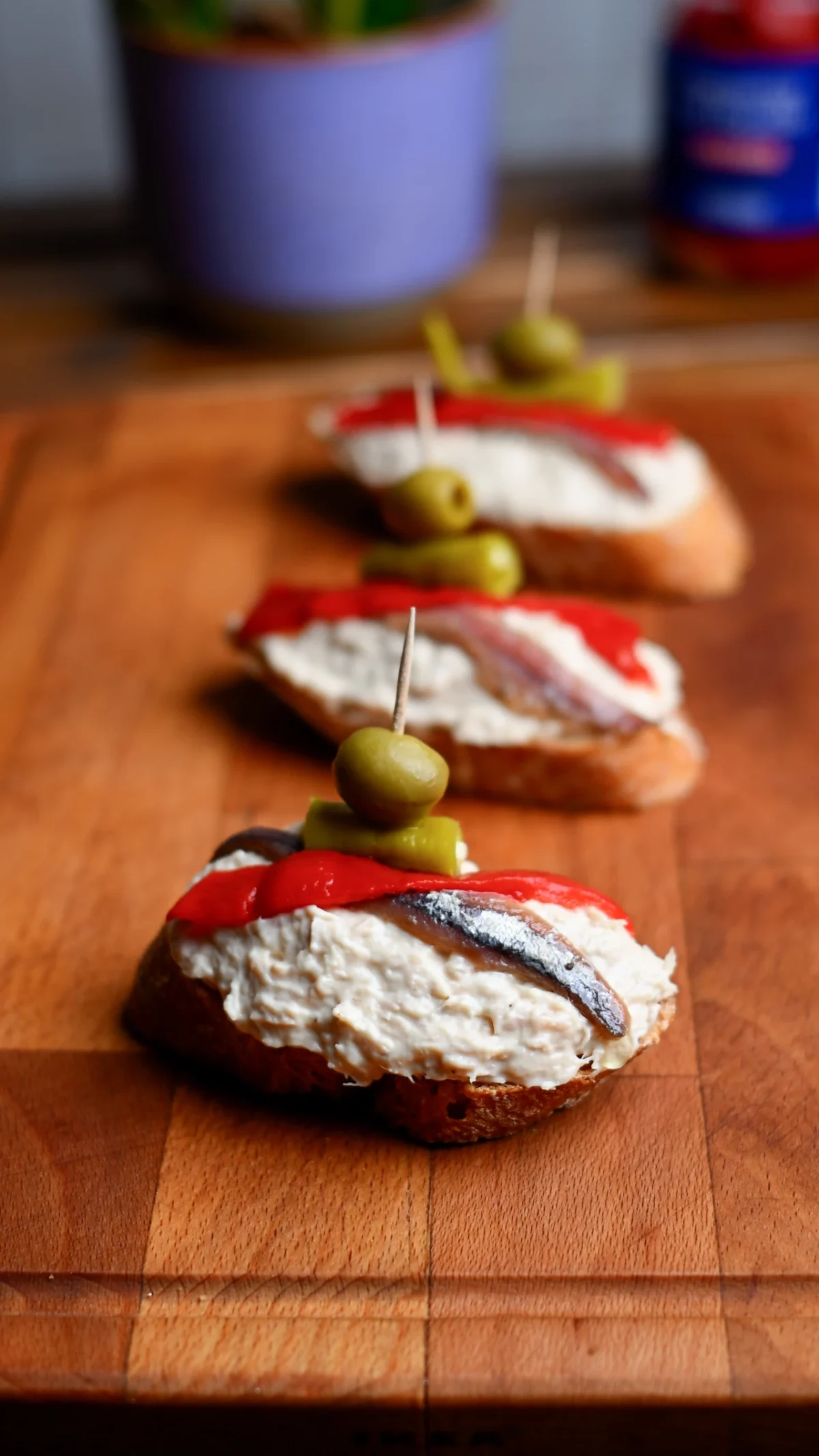 Pintxo de bonito — three baguette slices topped with tuna spread, roasted red pepper and anchovy, skewered with olives on a wooden board, Basque Country