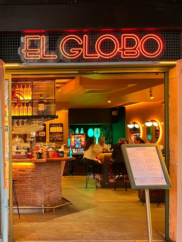 El Globo pintxo bar — red neon sign above an open entrance with a warm-lit interior, brick bar counter and menu board outside, Bilbao