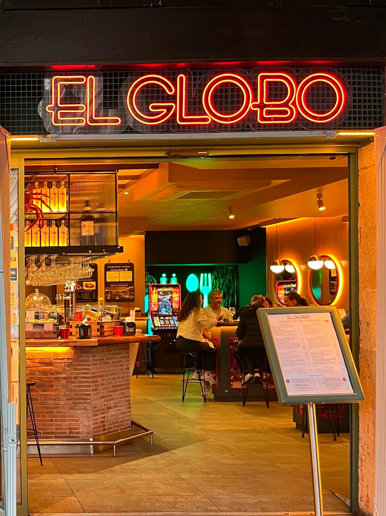 El Globo pintxo bar — red neon sign above an open entrance with a warm-lit interior, brick bar counter and menu board outside, Bilbao