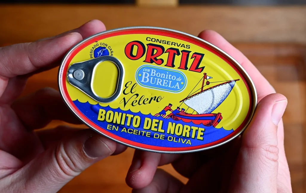 Pintxo de bonito recipe step 1 — hands holding an Ortiz Bonito del Norte in olive oil tin over a wooden board