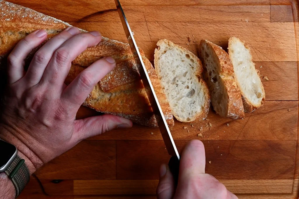 Pintxo de bonito recipe step 4 — hands slicing a crusty baguette into rounds on a wooden chopping board