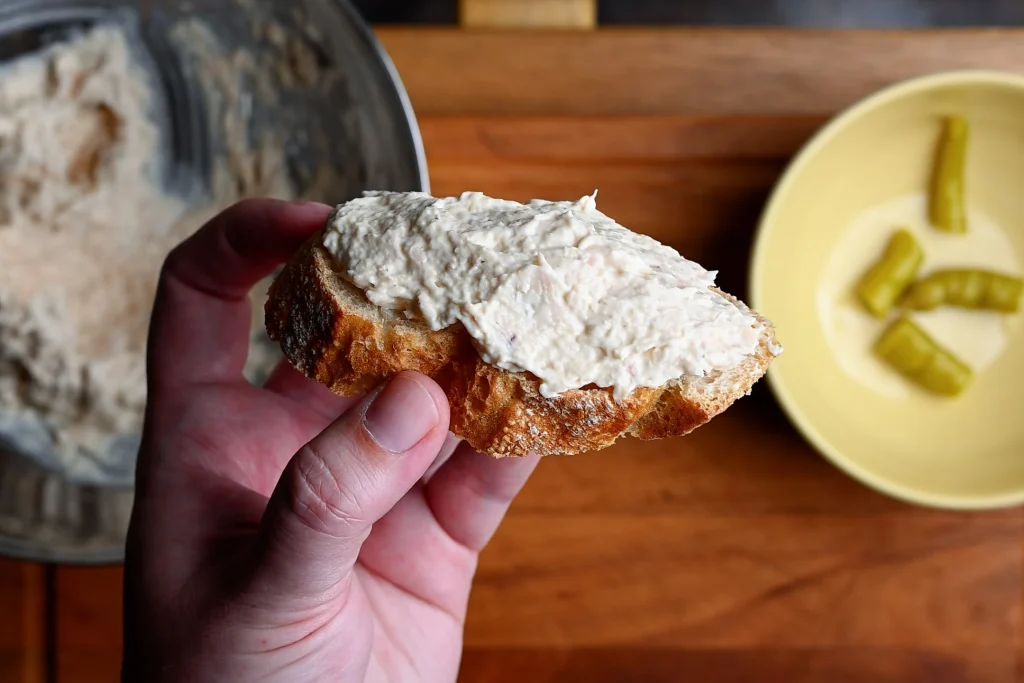 Pintxo de bonito recipe step 5 — hand holding a baguette slice spread with bonito and mayo paste, with guindillas and mixing bowl in background