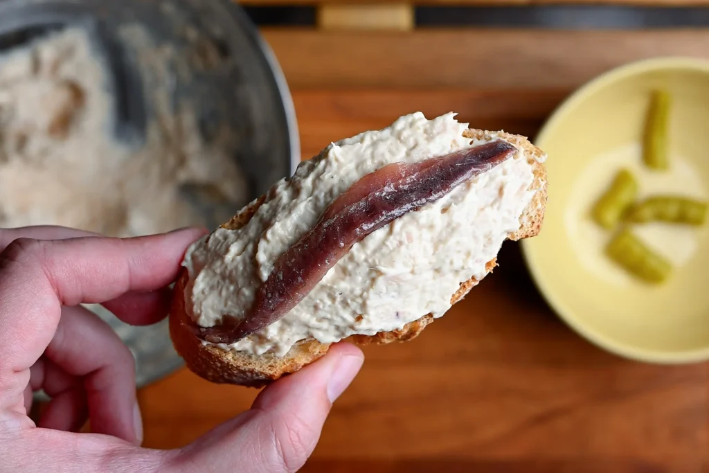 Pintxo de bonito recipe step 6 — a single anchovy fillet laid lengthways on bonito mayo spread on a baguette slice