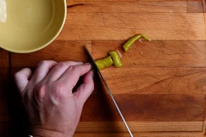 Pintxo de bonito recipe step 8 — hand cutting a guindilla pickled pepper into pieces on a wooden chopping board