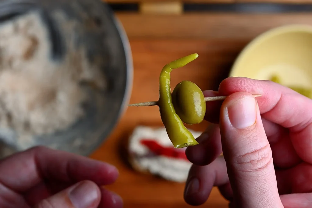 Pintxo de bonito recipe step 9 — hands threading a green olive and guindilla piece onto a toothpick held up close
