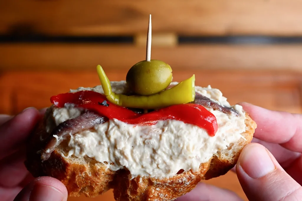 Pintxo de bonito recipe step 10 — hands holding a finished pintxo with toothpick, olive, guindilla, red pepper and anchovy on bonito spread
