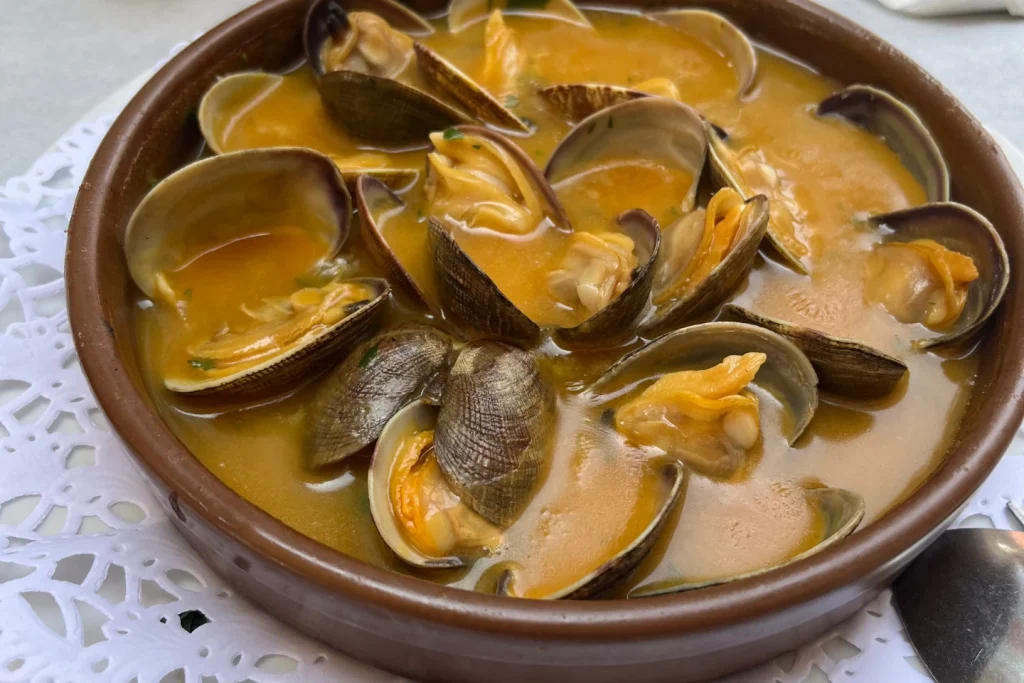Almejas a la marinera — open clams in a rich golden broth in a clay cazuela on a white lace doily, Spain