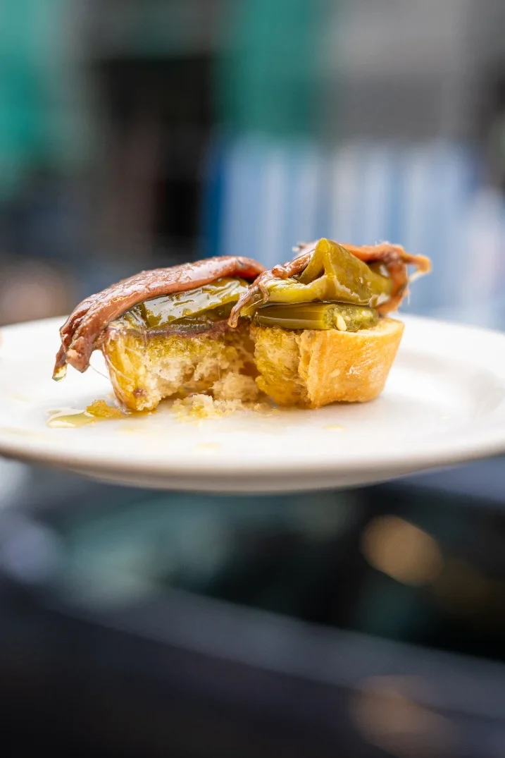Pintxo de antxoa — anchovy fillet and green pepper on an oil-soaked baguette slice held on a white plate at Antonio Bar, San Sebastián