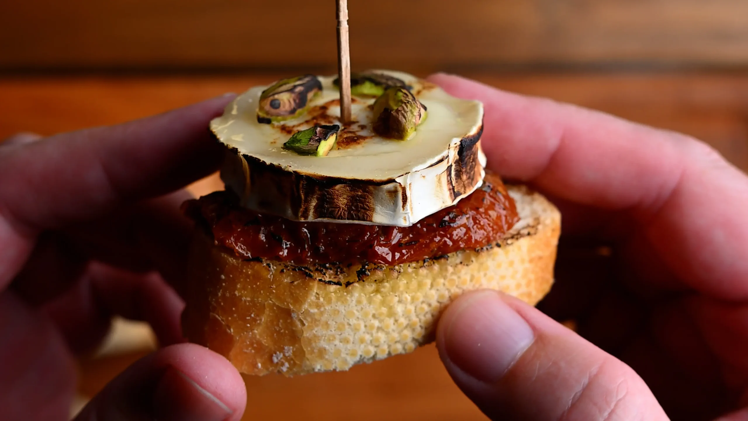 Pintxo de queso de cabra recipe — hands holding a baguette slice layered with dried tomato, charred goat's cheese and pistachio on a toothpick