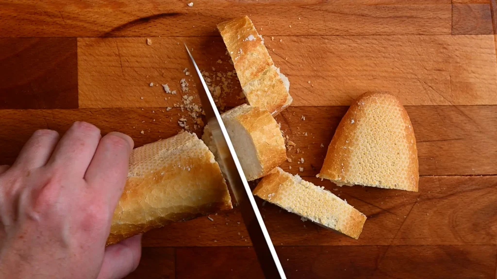 Pintxo de queso de cabra recipe step 1 — hands slicing a crusty baguette into rounds on a wooden chopping board