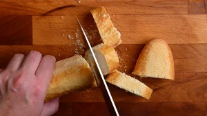 Pintxo de queso de cabra recipe step 1 — hands slicing a crusty baguette into rounds on a wooden chopping board