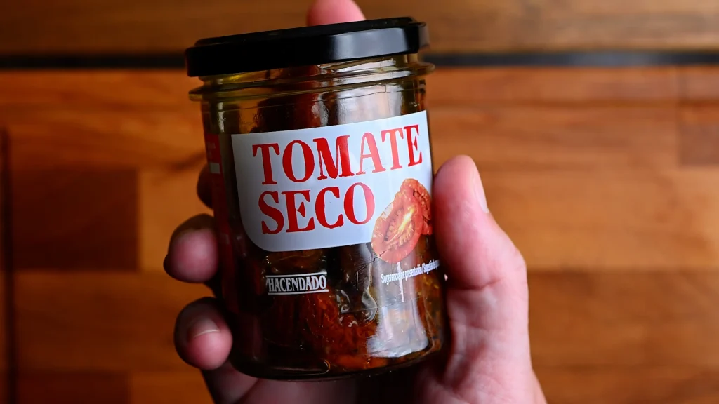 Pintxo de queso de cabra recipe step 2 — hand holding a Hacendado tomate seco jar of dried tomatoes in olive oil