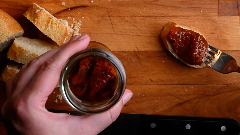 Pintxo de queso de cabra recipe step 3 — dried tomato placed on a baguette slice on a wooden board, jar and bread slices visible alongside