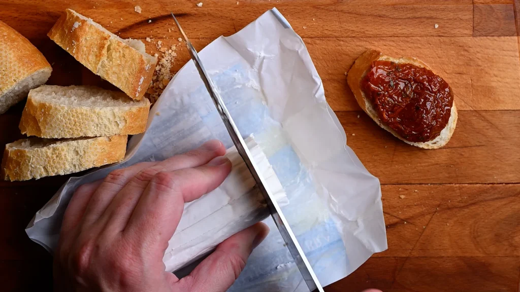 Pintxo de queso de cabra recipe step 4 — hand slicing a rolled goat's cheese log with a knife on paper, baguette slices and tomato bread in background