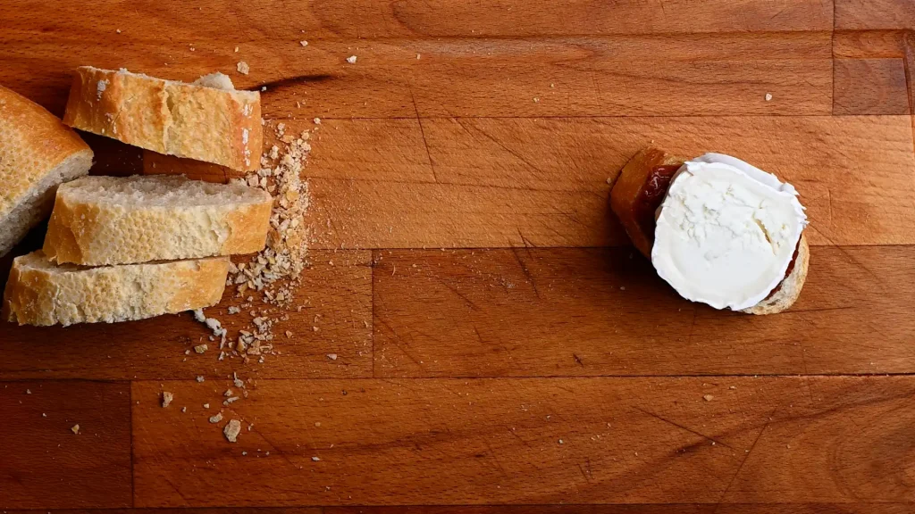 Pintxo de queso de cabra recipe step 5 — a goat's cheese slice placed on top of dried tomato on a baguette, overhead view on a wooden board