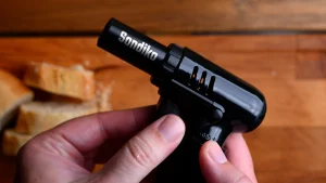 Pintxo de queso de cabra recipe step 6 — hands holding a Sondiko kitchen torch with baguette slices visible in the background