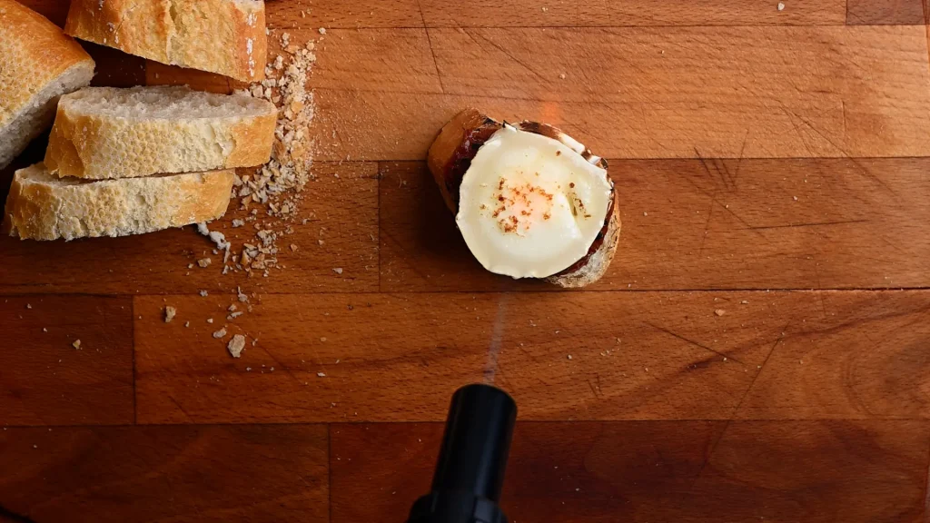Pintxo de queso de cabra recipe step 7 — kitchen torch aimed at a charred goat's cheese on baguette, overhead view on a wooden board