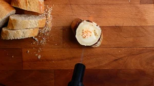 Pintxo de queso de cabra recipe step 7 — kitchen torch aimed at a charred goat's cheese on baguette, overhead view on a wooden board