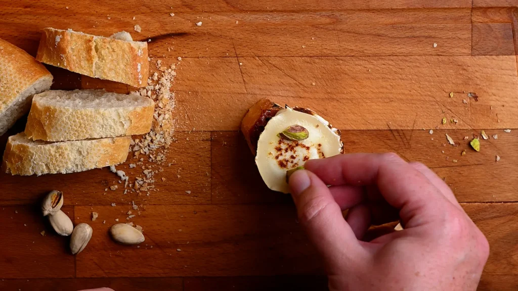 Pintxo de queso de cabra recipe step 8 — hand placing pistachios onto charred goat's cheese on baguette, whole pistachios visible on board
