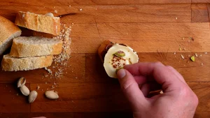 Pintxo de queso de cabra recipe step 8 — hand placing pistachios onto charred goat's cheese on baguette, whole pistachios visible on board