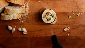 Pintxo de queso de cabra recipe step 9 — kitchen torch aimed at pistachios on goat's cheese and baguette, overhead view on a wooden board