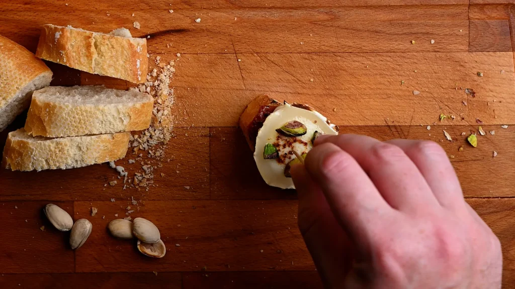 Pintxo de queso de cabra recipe step 10 — hand pressing a toothpick through charred goat's cheese and pistachios into the baguette on a wooden board