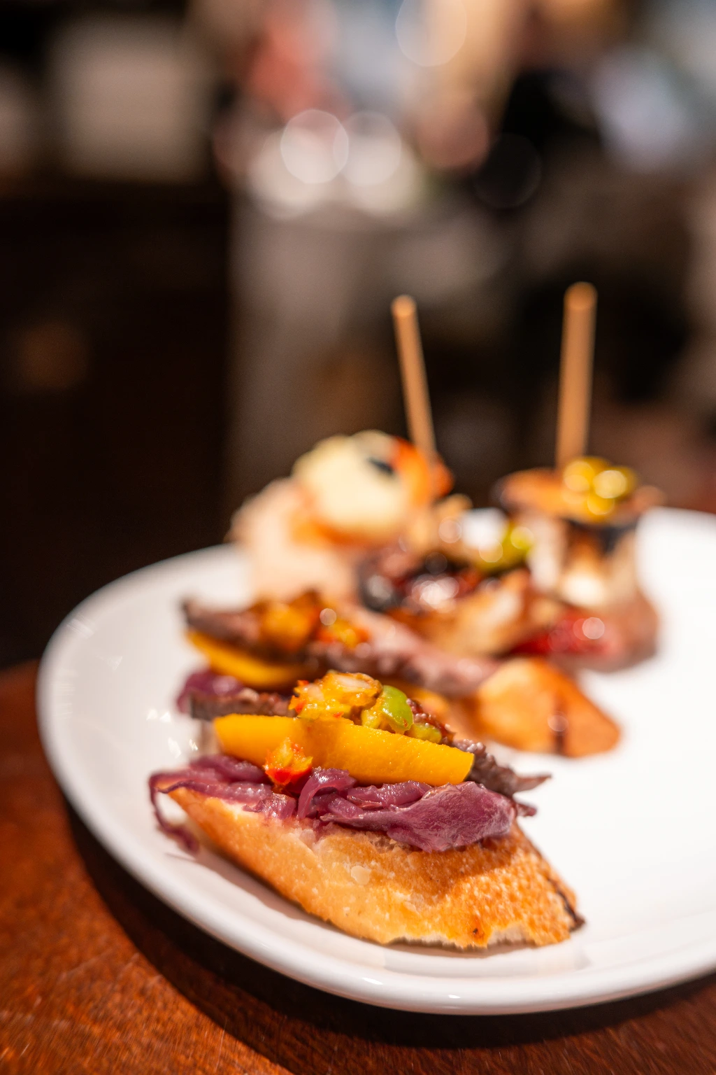 A pintxo on a baguette slice topped with braised meat and roasted yellow and green peppers on a white plate, San Sebastián
