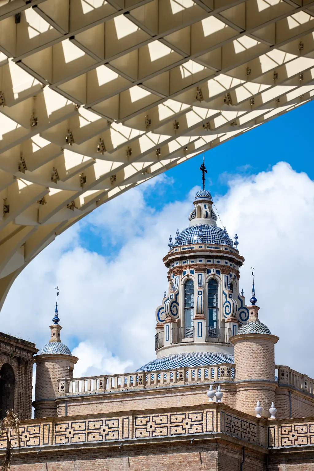 The tiled dome of Plaza de España framed by the underside of Las Setas wooden structure against a blue sky, Seville