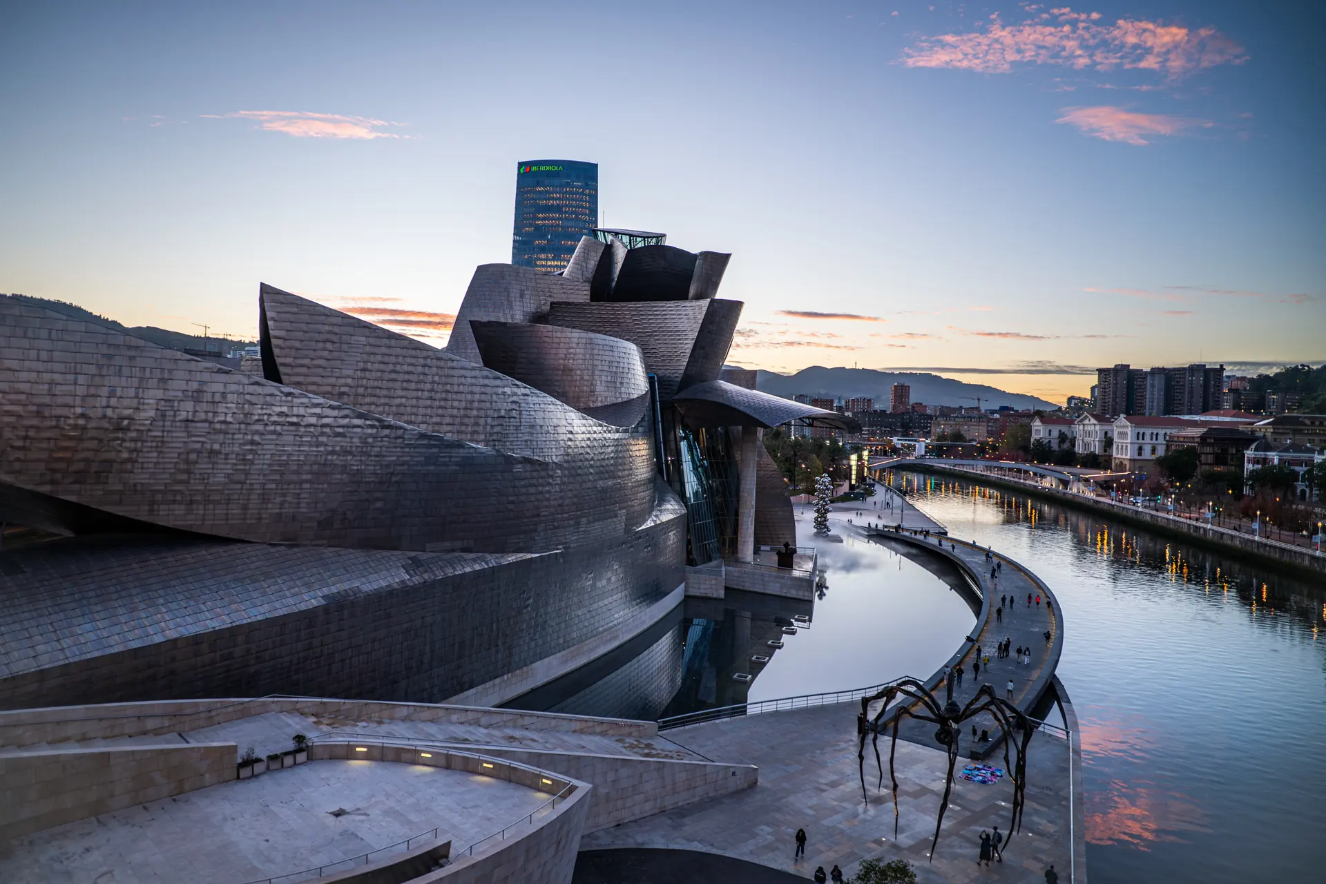 Guggenheim Museum Bilbao titanium facade with Maman spider sculpture and Nervión river at dusk, Bilbao, Spain