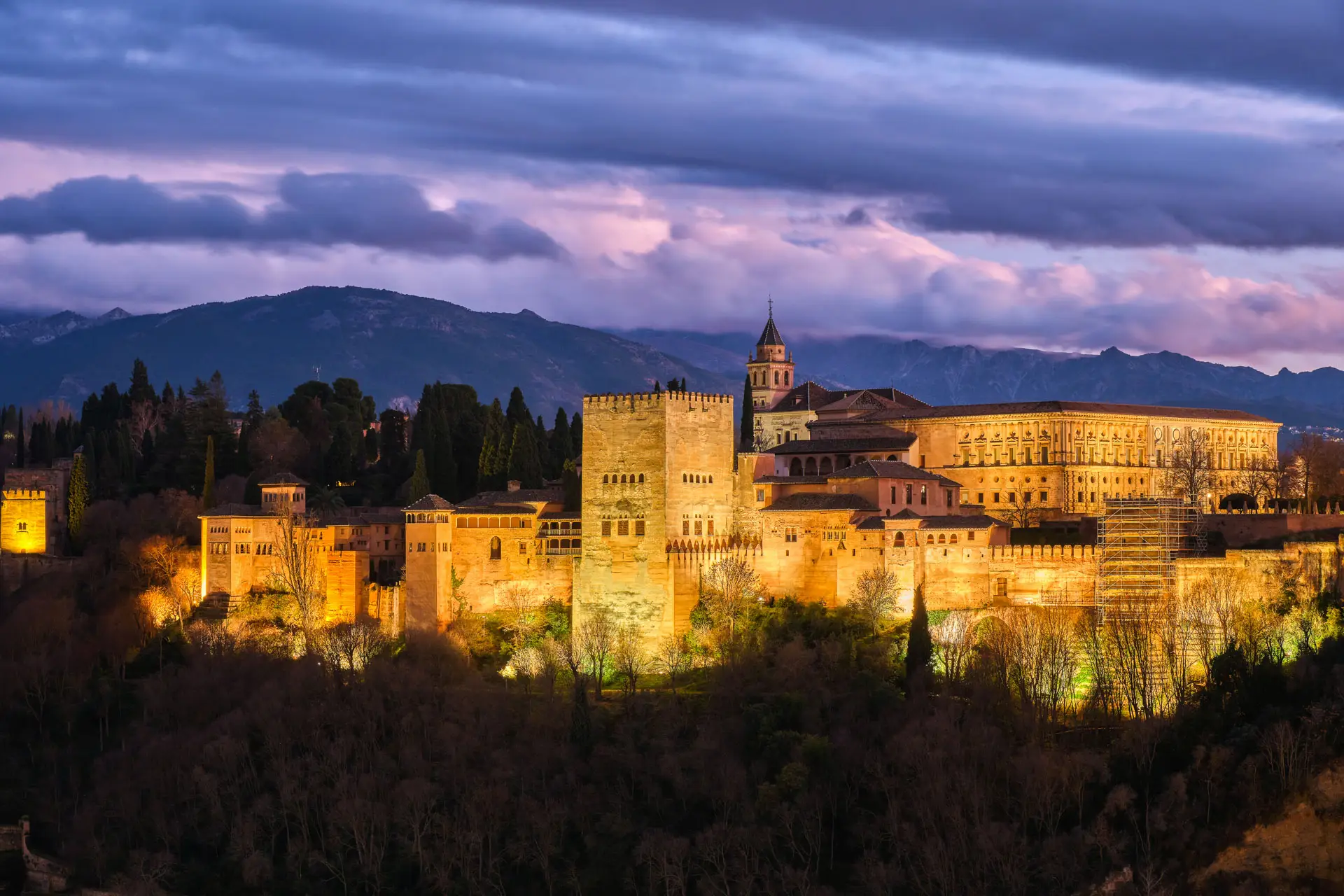 Alhambra palace complex illuminated at dusk with Sierra Nevada mountains under a purple sky, Granada, Spain
