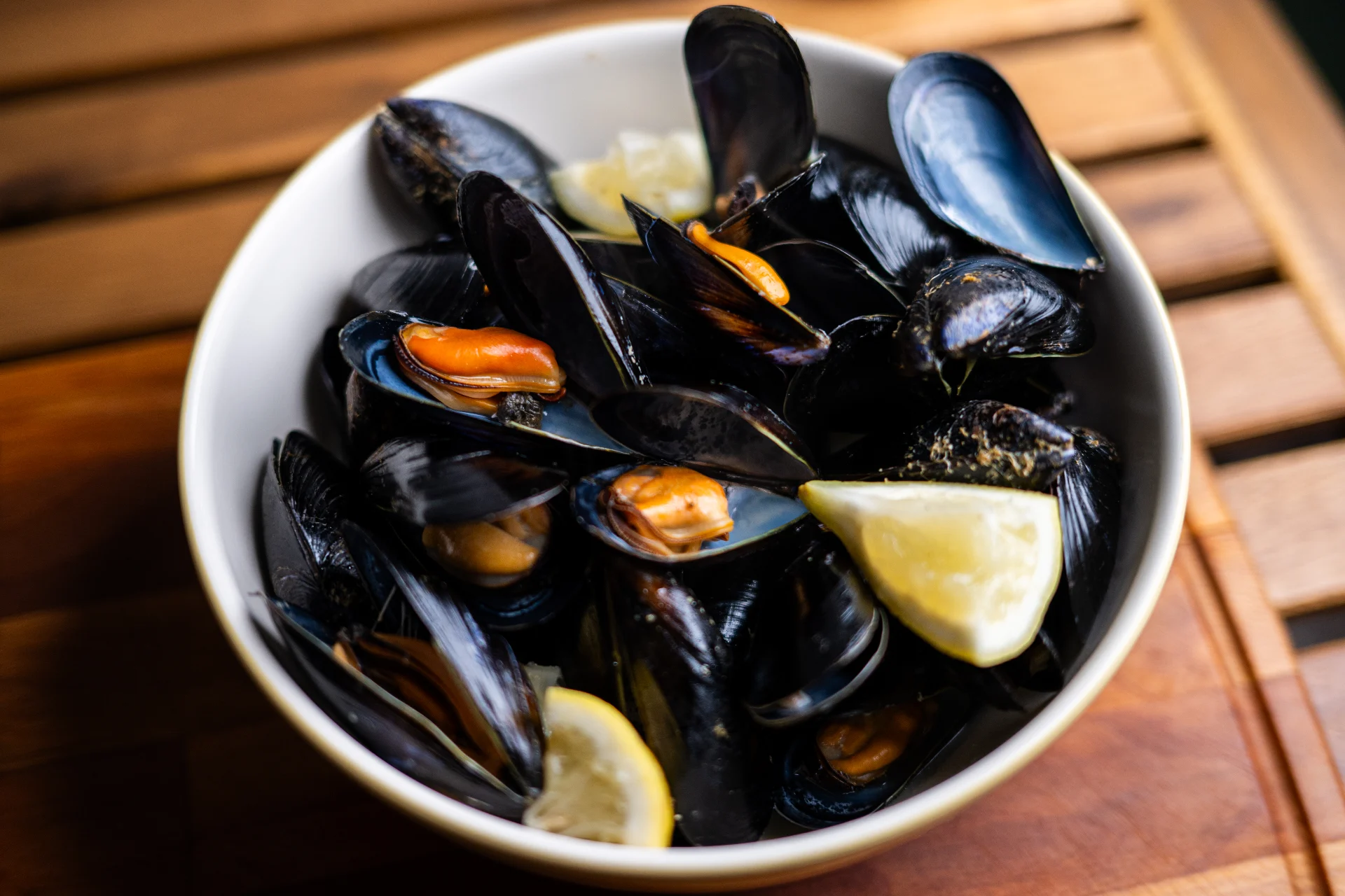 Mejillones al vapor — steamed mussels with orange flesh in a dark-rimmed bowl, two lemon wedges, on a slatted wooden table