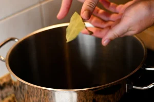 Mejillones al vapor step 2 — hands crushing a bay leaf over an empty stainless steel pot on a ceramic hob