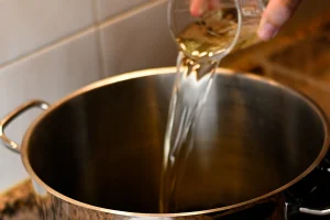 Mejillones al vapor step 4 — a hand pouring white wine in a wide stream into a large stainless steel pot on a hob