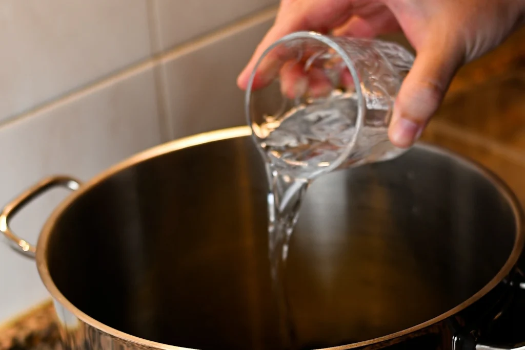 Mejillones al vapor step 5 — a hand pouring water from a glass into a large stainless steel pot on a ceramic hob