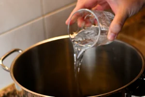 Mejillones al vapor step 5 — a hand pouring water from a glass into a large stainless steel pot on a ceramic hob