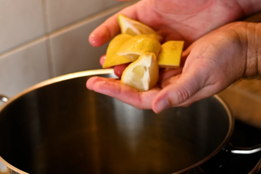 Mejillones al vapor step 6 — hands holding lemon wedges over a stainless steel pot on a hob, about to add them to the steam