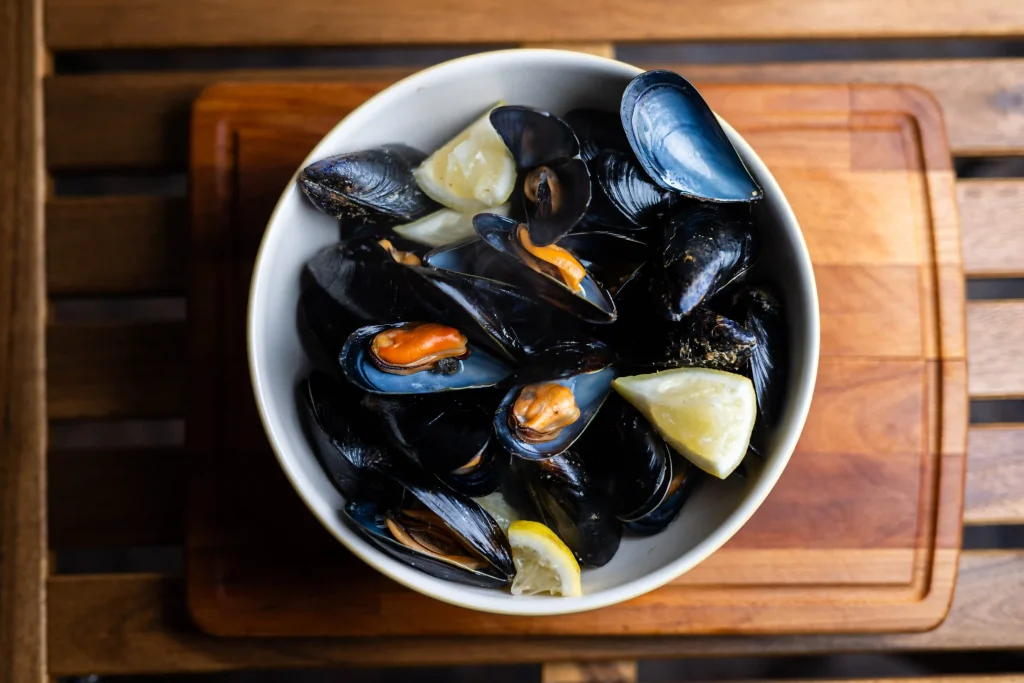 Mejillones al vapor step 8 — steamed mussels in a white bowl on a wooden chopping board, two lemon wedges, served hot