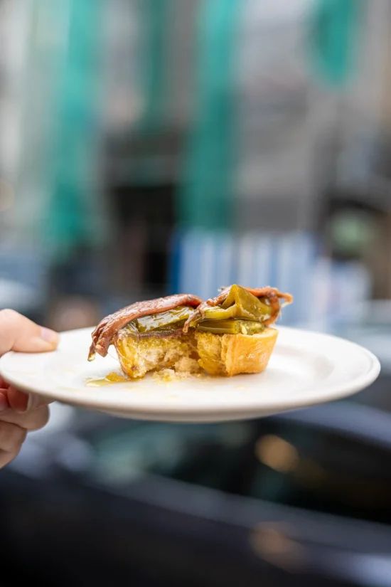 Pintxo de antxoa — anchovy fillet and green pepper on an oil-soaked baguette slice held on a white plate at Antonio Bar, San Sebastián