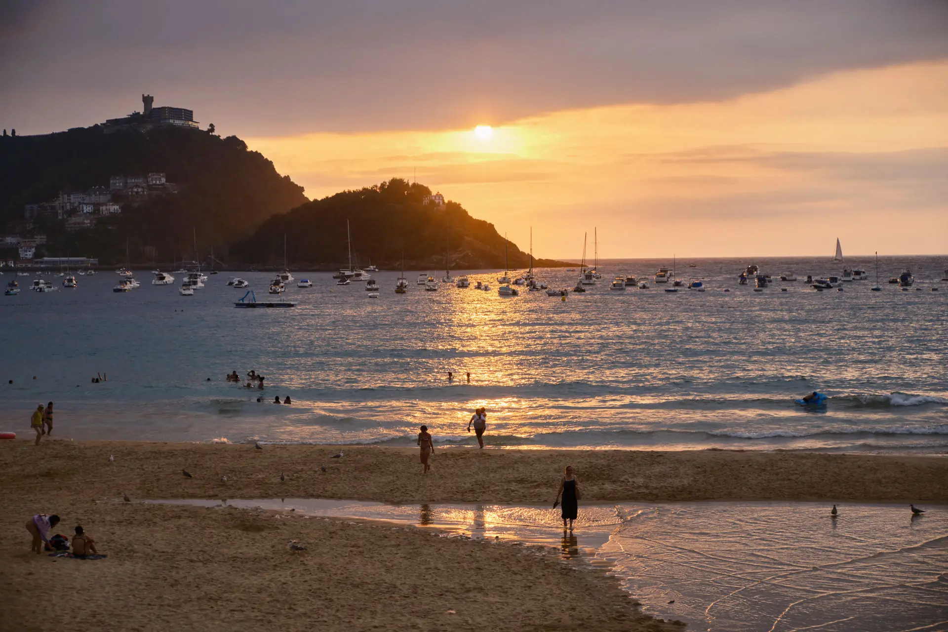 La Concha beach at sunset with silhouetted figures, anchored sailboats and Monte Igueldo in the background, San Sebastián, Spain