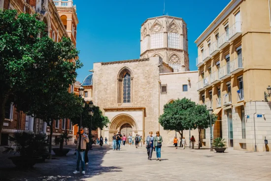 Valencia Cathedral Gothic facade on Plaza de la Reina with orange trees and people walking under a blue sky, Valencia, Spain