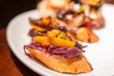 A pintxo on a baguette slice topped with braised meat and roasted yellow and green peppers on a white plate, San Sebastián