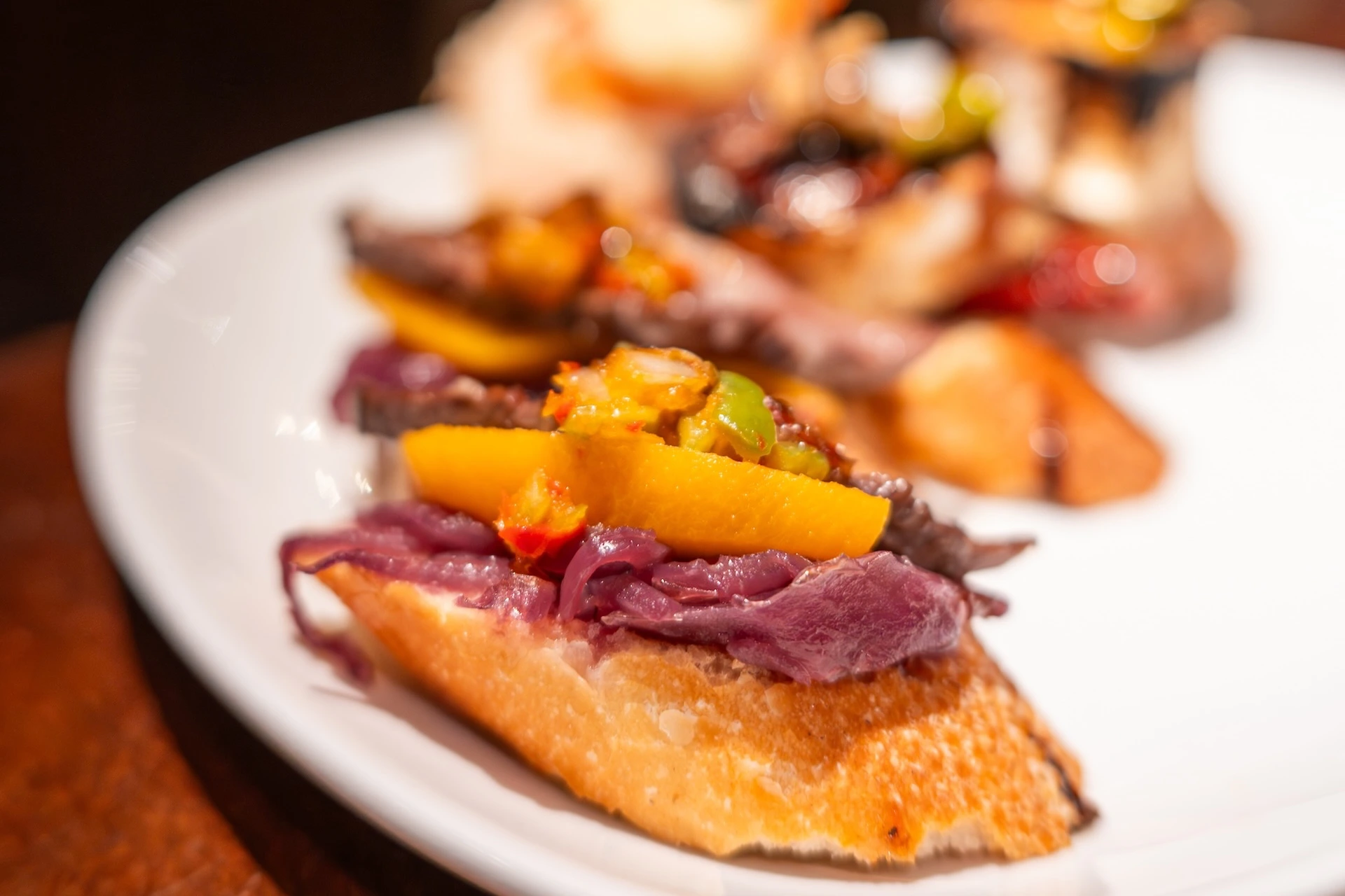 A pintxo on a baguette slice topped with braised meat and roasted yellow and green peppers on a white plate, San Sebastián