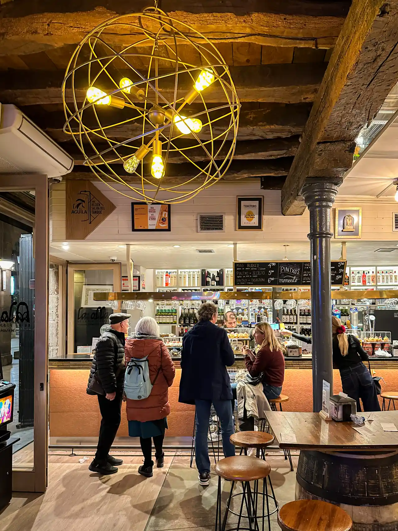 Spanish pintxos bar interior — customers standing at a lit counter, globe pendant light, exposed timber ceiling and chalkboard menu