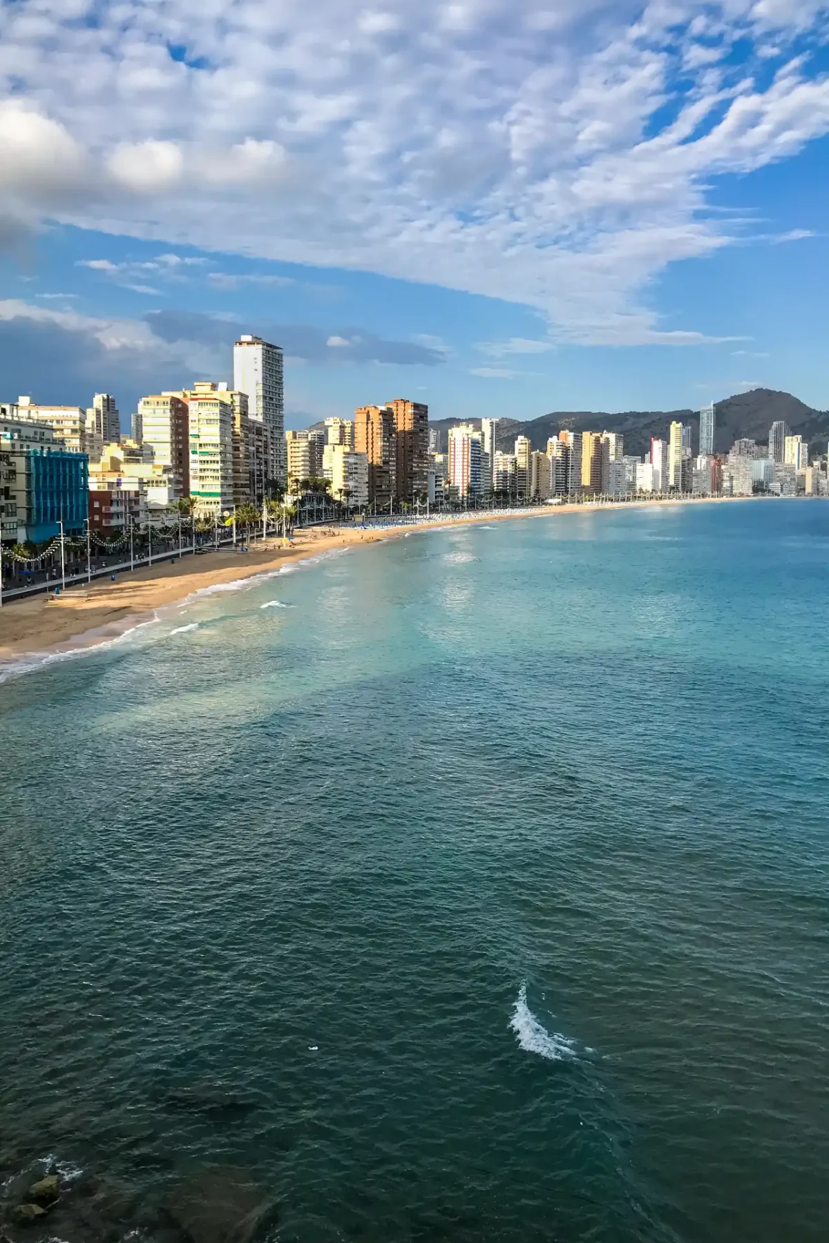 Benidorm Levante beach — long arc of sand backed by a dense high-rise skyline and turquoise sea, with mountains behind, Alicante
