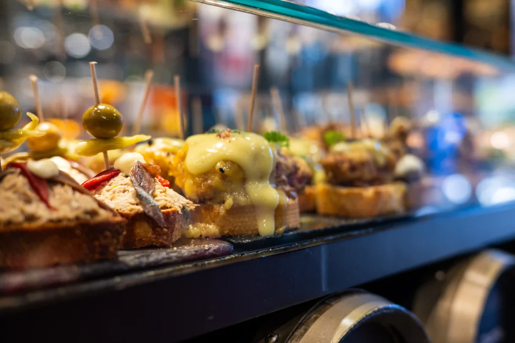 Spanish pintxos counter — rows of topped bread slices with anchovies, melted cheese, olive skewers and peppers under a glass screen