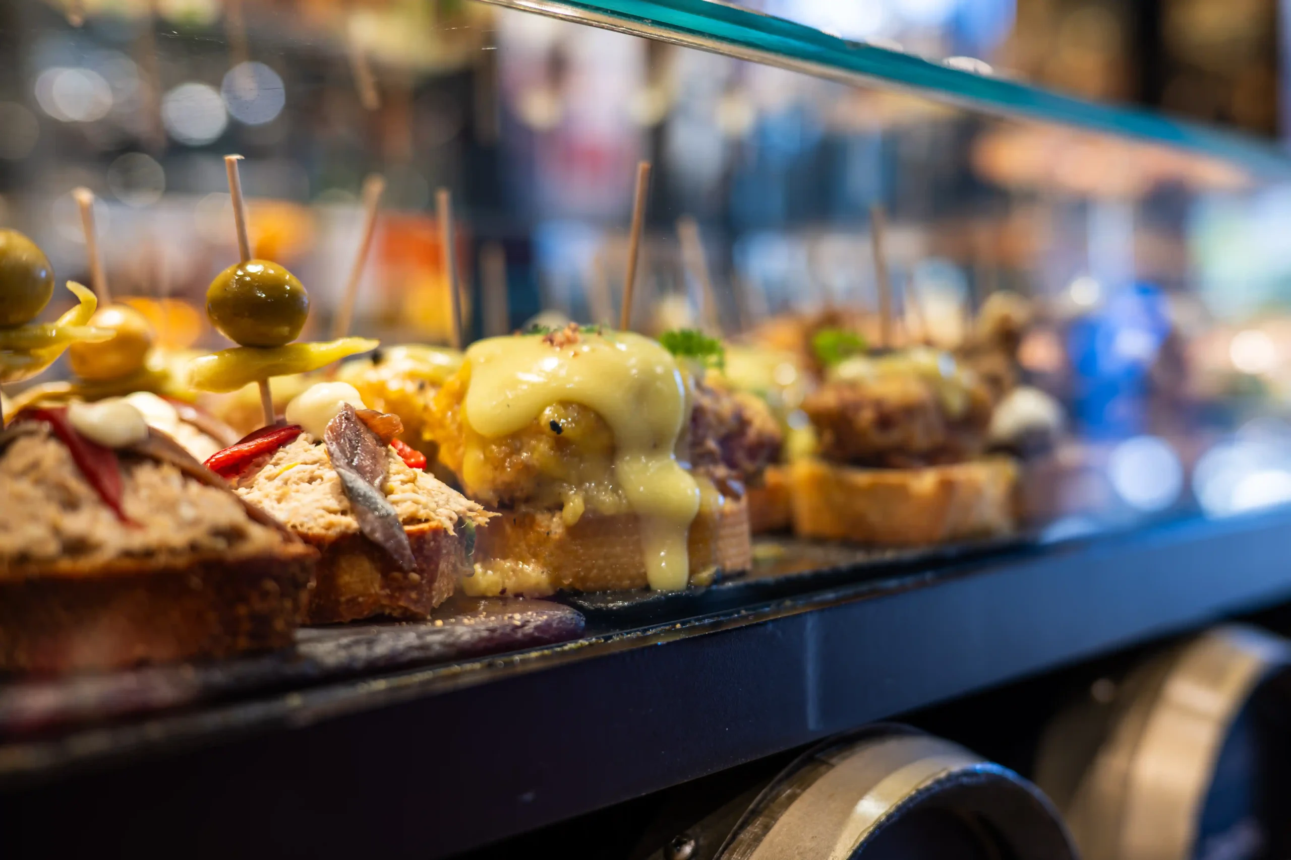 Spanish pintxos counter — rows of topped bread slices with anchovies, melted cheese, olive skewers and peppers under a glass screen