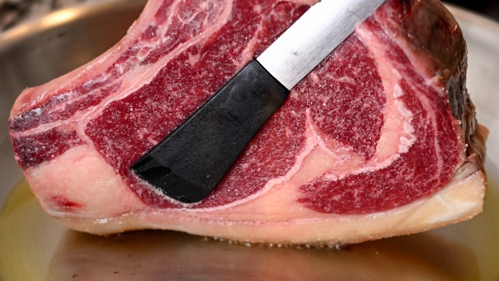 Holding a seasoned chuleton steak sideways with tongs to render the fat edge in a hot pan, Spain.